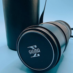 May include: A black camera lens with the brand name "SIGMA" in white lettering on the lens cap. The lens is lying next to a black cylindrical case with a strap. The background is a light blue.