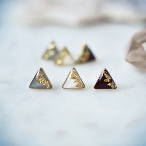 May include: Three pairs of triangle-shaped earrings with gold leaf accents. The earrings are in gray, white, and black.