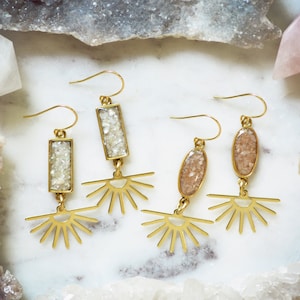 May include: A set of three pairs of gold earrings with a sunburst design. Each earring features a rectangular or oval shaped stone with a white or pink speckled pattern.