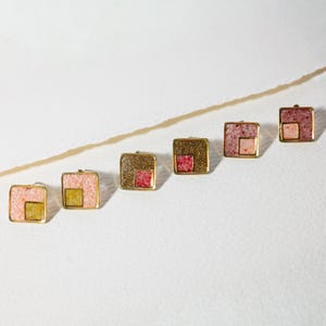 May include: Six square earrings with gold-coloured frames. Each earring features a textured surface with a smaller square in the centre. The centre squares vary in colour, including pink, yellow, and red.