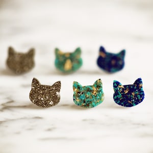 May include: Six cat-shaped resin earrings with glitter embedded in the resin. The earrings are in various colors, including black, gold, blue, and green.