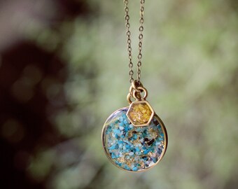 May include: A gold chain necklace with a round pendant filled with blue and gold glitter. The pendant is attached to a smaller hexagonal pendant filled with gold glitter.