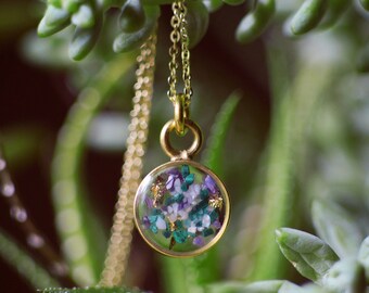May include: A gold chain necklace with a round pendant featuring a clear resin filled with purple, green, and white flowers and gold flakes.