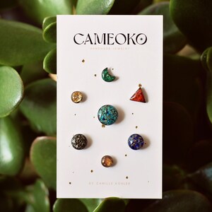 May include: A set of seven handmade resin earrings in various shapes and colors, including a crescent moon, a triangle, a circle, and a star. The earrings are displayed on a white card with the text "Cameoko Handmade Jewelry" and "By Camille Kohler".