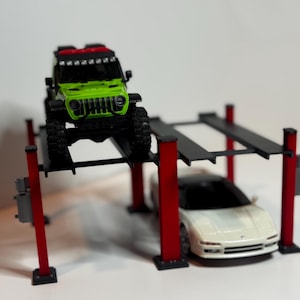 May include: A model car lift featuring a green Jeep on the upper platform and a white sports car below. The lift has red supports and black platforms. The Jeep has black tyres and a black grille.