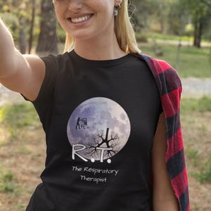 May include: Black t-shirt featuring a large, full moon design with a silhouette of a person and tree branches. The text "R.T. The Respiratory Therapist" is printed below the moon. The person is wearing a blue cap.