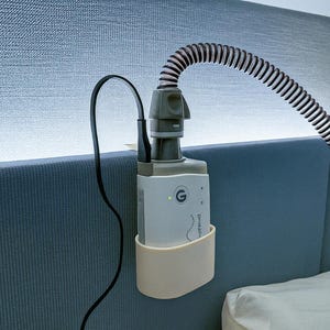 Könnte beinhalten: Ein weißes und graues medizinisches Gerät mit einem schwarz-weiß gestreiften Schlauch ist an einem blauen Kopfteil befestigt. Ein schwarzes Netzkabel ist mit dem Gerät verbunden. Das Gerät hat ein kleines grünes Licht und ein geschwungenes Design.