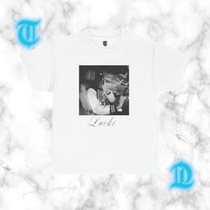 Könnte beinhalten: Ein weißes kurzärmeliges T-Shirt mit einer quadratischen Schwarz-Weiß-Grafik einer Person in einer College-Jacke. Das Wort Lucki ist in einer Schreibschrift unter der Grafik gedruckt. Ein kleiner gotischer Buchstabe T befindet sich am Innenetikett.