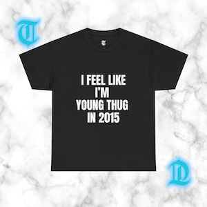 May include: A black short-sleeved t-shirt featuring the white text I FEEL LIKE I'M YOUNG THUG IN 2015 printed in the center of the chest. The garment has a crew neckline and a standard fit, showcasing a clean and simple design on a solid black fabric.