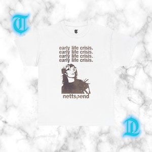 May include: A white short-sleeved t-shirt featuring a dark brown graphic print. The design includes the repeated text "early life crisis." above a stylized portrait of a person, with the word "nettspend" printed in lowercase letters below.