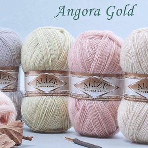 May include: A selection of Angora Gold yarn skeins in shades of pink, cream, and grey. Each ball is labelled with "ALIZE Angora Gold." A crochet hook and a dried leaf are in the foreground. The text "Angora Gold" is at the top.