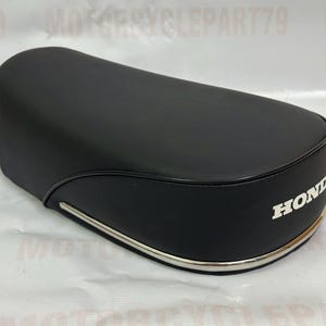 Può includere: Sella per moto nera di forma arrotondata con finiture argentate. La scritta "HONDA" è stampata in bianco sul lato. La sella è progettata per una moto.