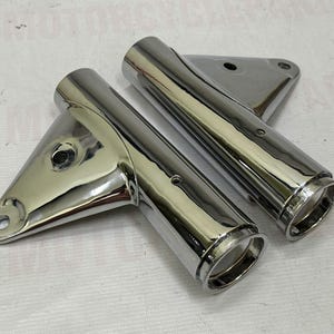 May include: Two chrome motorcycle headlight brackets. The brackets are highly polished and reflect the surrounding environment. Each bracket has a cylindrical section and a mounting plate with a hole for attachment. The parts are new and shiny.