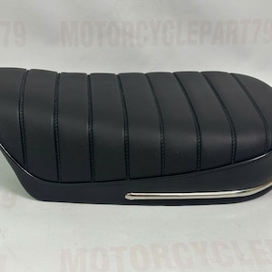 Può includere: Sella da moto nera con design a coste. Il sedile ha una forma curva e un rivestimento cromato lungo il bordo inferiore. Il sedile è realizzato in un materiale liscio simile alla pelle.