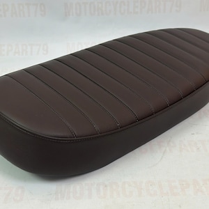 Peut inclure: Une selle de moto marron avec un design c&ocirc;tel&eacute;. Le si&egrave;ge est de forme ovale et a une texture lisse semblable au cuir. Les coutures sont visibles. La selle semble neuve.