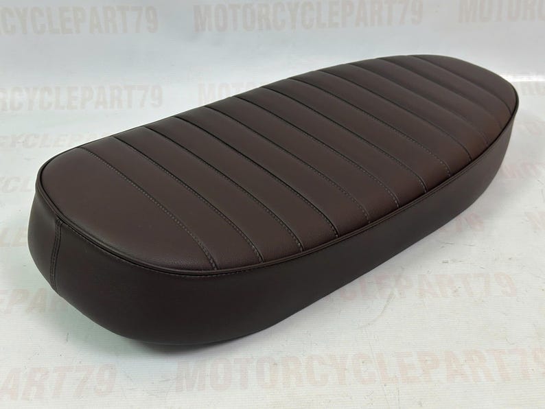 Peut inclure: Une selle de moto marron avec un design c&ocirc;tel&eacute;. Le si&egrave;ge est de forme ovale et a une texture lisse semblable au cuir. Les coutures sont visibles. La selle semble neuve.