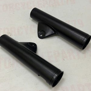 May include: Two black motorcycle fork tubes with mounting brackets. The tubes are cylindrical with a smooth, matte finish. Each tube has a flared end. The brackets have a single mounting hole.