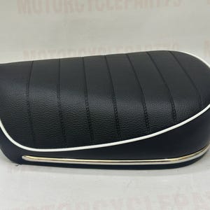 May include: Black motorcycle seat with vertical stitching and a white trim. The seat has a textured surface and a gold-coloured metal trim along the bottom edge. The seat is designed for a motorcycle.