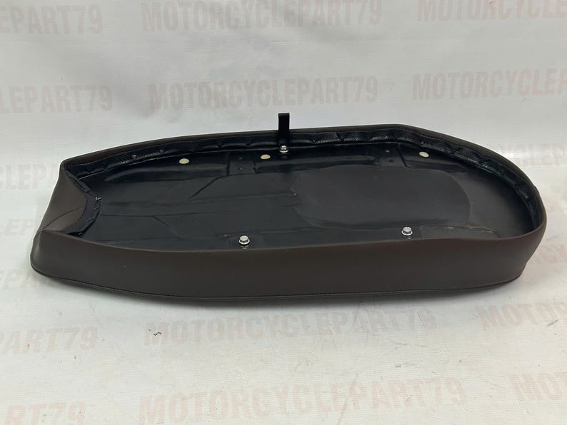 Peut inclure: Une selle de moto marron avec un int&eacute;rieur noir. La selle a une forme arrondie et est fix&eacute;e avec plusieurs fixations m&eacute;talliques. Un petit support noir est fix&eacute; sur le dessus de la selle.