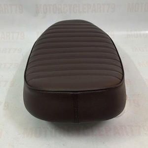 Peut inclure: Une selle de moto marron avec une surface rembourr&eacute;e et c&ocirc;tel&eacute;e. Le si&egrave;ge a une forme arrondie et est fait d'un mat&eacute;riau lisse semblable au cuir. Le si&egrave;ge est con&ccedil;u pour une moto et est une pi&egrave;ce de rechange.