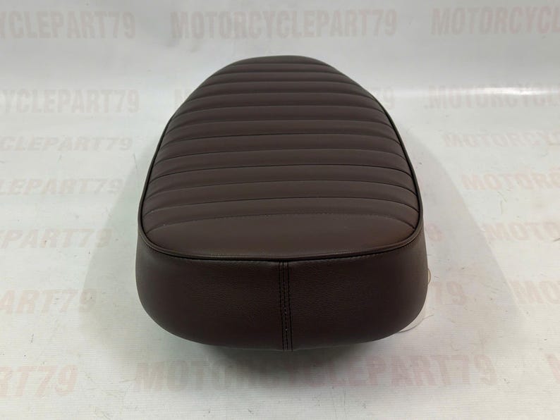 Peut inclure: Une selle de moto marron avec une surface rembourr&eacute;e et c&ocirc;tel&eacute;e. Le si&egrave;ge a une forme arrondie et est fait d'un mat&eacute;riau lisse semblable au cuir. Le si&egrave;ge est con&ccedil;u pour une moto et est une pi&egrave;ce de rechange.