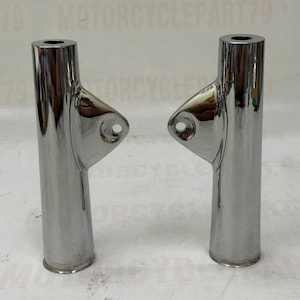 May include: Two chrome motorcycle fork tubes. Each tube has a cylindrical body with a flared base and a small, angled bracket with a hole. The tubes are highly reflective, with a polished finish.