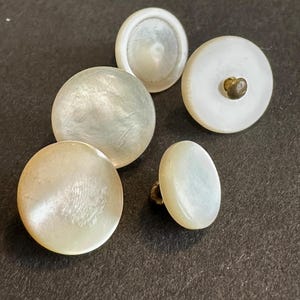 May include: Five vintage buttons of varying sizes and shapes. The buttons are mainly white with a pearlescent sheen, and some have an iridescent quality. One button features a brass shank.