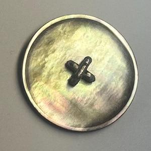 May include: A round, iridescent button with a cross-shaped center. The button has a mother-of-pearl appearance with a dark, metallic cross in the center. The button is likely for clothing or crafts.