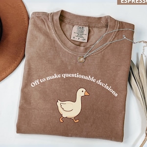 Puede incluir: Camiseta color espresso con la frase "Off to make questionable decisions" y un dibujo de un pato. Un collar plateado con un colgante descansa sobre la camiseta. Marca Comfort Colors.