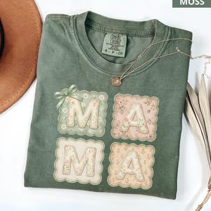Puede incluir: Una camiseta verde musgo con la palabra "MAMA" en cuadrados con estampado floral. La camiseta tiene cuello redondo y mangas cortas. Un collar dorado con un colgante está drapeado sobre la camiseta. Un sombrero marrón y una planta seca están en el fondo.