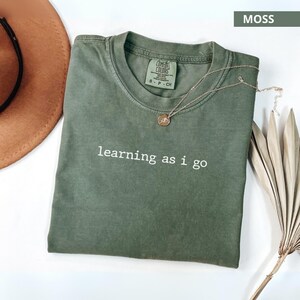 Puede incluir: Una camiseta verde musgo con la frase "learning as I go" impresa en blanco. Un collar dorado con un colgante descansa sobre la camiseta. Un sombrero marrón y una planta seca también están en la imagen.