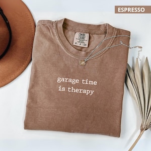 Puede incluir: Camiseta color espresso con el texto "garage time is therapy" en blanco. La camiseta tiene cuello redondo y una pequeña etiqueta. También se ve un collar plateado con un colgante y un sombrero marrón.