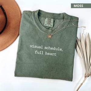 Puede incluir: Una camiseta verde musgo con el texto "visual schedule, full heart" en blanco. Un collar dorado con un colgante y un sombrero marrón están junto a la camiseta. La camiseta es de un material suave.