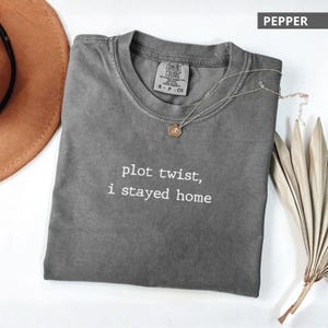 Puede incluir: Una camiseta gris Comfort Colors con el texto "plot twist, i stayed home" en blanco. Un collar dorado con un colgante descansa sobre la camiseta. La camiseta está doblada y un sombrero marrón es parcialmente visible en el fondo.