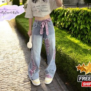 Könnte beinhalten: Blaue Jeans aus Denim mit pink-weißem kariertem Patchwork-Detail. Die Jeans hat eine hohe Taille und eine weite Beinweite. Das Model trägt ein weißes Crop-Top mit dem Text "Pink soft sisters are the funkiest" und "0 Minute".