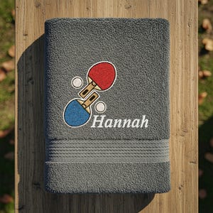Pode incluir: Toalha de banho cinza bordada com um design de raquetes de pingue-pongue vermelhas e azuis e o nome "Hannah" em branco. A toalha tem uma superfície texturizada e uma faixa decorativa na parte inferior.