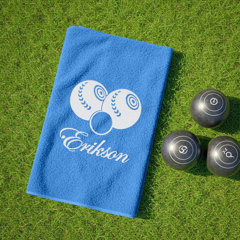 Puede incluir: Una toalla azul con un dise&ntilde;o de bolas de bolos blancas y el nombre "Erikson". Tres bolas de bolos negras con marcas blancas est&aacute;n sobre hierba verde. La toalla es rectangular y est&aacute; doblada.