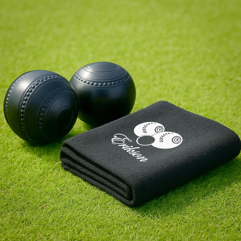 Puede incluir: Dos bolas de c&eacute;sped negras con ranuras decorativas y una toalla negra con el nombre "Erickson" y un gr&aacute;fico blanco. Los art&iacute;culos est&aacute;n colocados sobre una superficie de hierba verde.