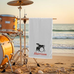 Può includere: Un telo da mare bianco con un disegno di batteria nera e il nome "Harrison" in rosso. Una batteria e piatti sono sullo sfondo su una spiaggia sabbiosa con conchiglie.