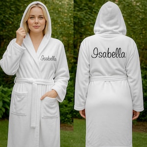 Könnte beinhalten: Ein weißer Bademantel mit Kapuze und dem Namen "Isabella" auf der Vorder- und Rückseite gestickt. Der Bademantel hat einen Gürtel, Taschen und eine weiche, plüschige Textur. Das Model trägt den Bademantel.