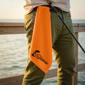 Puede incluir: Una toalla de pesca naranja con el nombre "Victoria" en letra cursiva y un gráfico de pez. La toalla cuelga del cinturón de una persona, junto a una caña de pescar. El fondo muestra un muelle de madera y el mar.