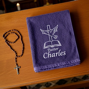 Op de afbeelding: Een paarse handdoek met witte borduursels en een duivenontwerp, naast een rozenkrans. De handdoek leest "Pastor Charles" onder een afbeelding van een duif boven een open boek en een kruis.