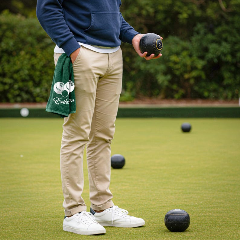 Puede incluir: Una persona de pie en un c&eacute;sped verde, sosteniendo una bola de c&eacute;sped negra y una toalla verde. La toalla tiene un gr&aacute;fico blanco y el nombre "Eriks" en blanco. La persona lleva pantalones color caqui, zapatillas blancas y una sudadera con capucha azul marino.