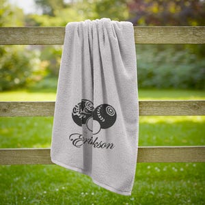 Puede incluir: Una toalla gris claro con un dise&ntilde;o decorativo de bolas de bolos y el nombre "Erikson" en cursiva. La toalla est&aacute; colgada de una valla de madera, con hierba verde en el fondo.