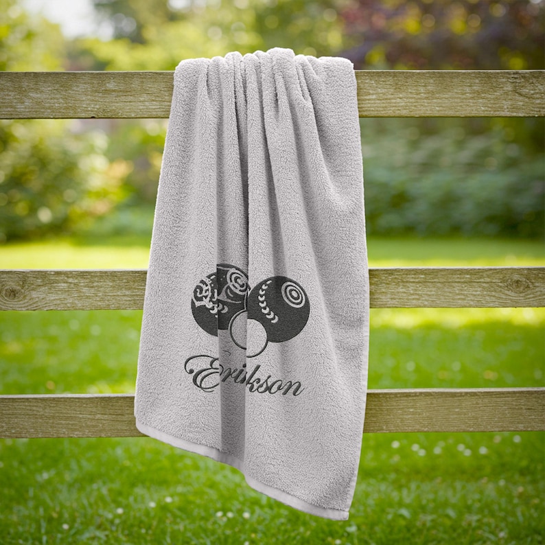 Puede incluir: Una toalla gris claro con un dise&ntilde;o decorativo de bolas de bolos y el nombre "Erikson" en cursiva. La toalla est&aacute; colgada de una valla de madera, con hierba verde en el fondo.