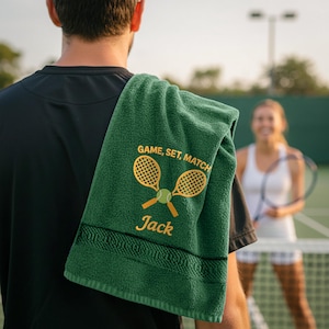 Peut inclure: Une serviette de tennis vert foncé avec les mots "GAME, SET, MATCH" et le nom "Jack" brodés en or. La serviette présente deux raquettes de tennis et une balle de tennis. La serviette est drapée sur l'épaule d'une personne.