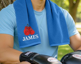 以下が含まれることがあります： 赤いボクシンググローブと「JAMES」の白い刺繍が入った青いスポーツタオル。タオルは首にかけられています。黒いボクシンググローブも確認できます。