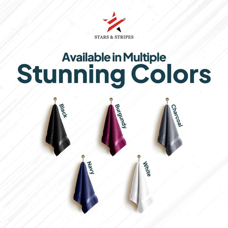 May include: Five towels hanging on hooks, showcasing different colors: black, burgundy, charcoal, navy, and white. The text "Available in Multiple Stunning Colors" is displayed above the towels.
