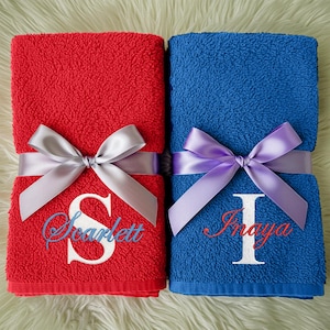 May include: Two folded towels, one red and one blue, are tied with satin ribbons. The red towel has the letter "S" and the name "Scarlett" embroidered in white and blue. The blue towel has the letter "I" and the name "Inaya" embroidered in white and red.