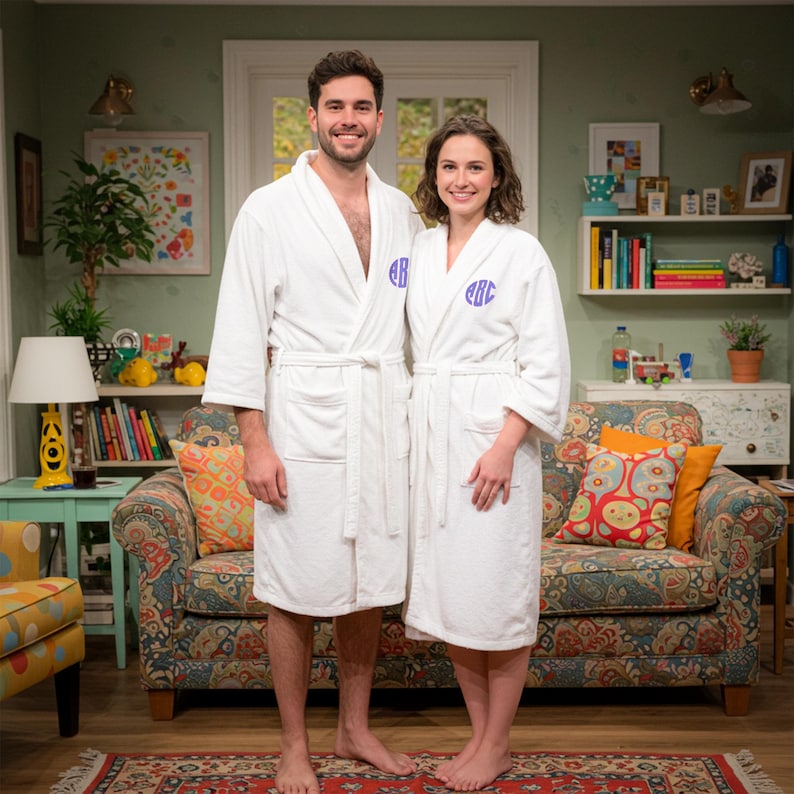 Puede incluir: Una pareja est&aacute; junta, ambos visten batas de ba&ntilde;o blancas de rizo. Cada bata tiene un monograma en hilo azul en el pecho. El fondo muestra una sala de estar con un sof&aacute; estampado, una alfombra y varios objetos decorativos.